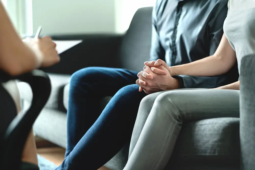 Couple sitting close together holding hands during a therapy session while speaking with a counselor - Finding Peace Counseling Service PLLC Couple sitting close together holding hands during a therapy session while speaking with a counselor.