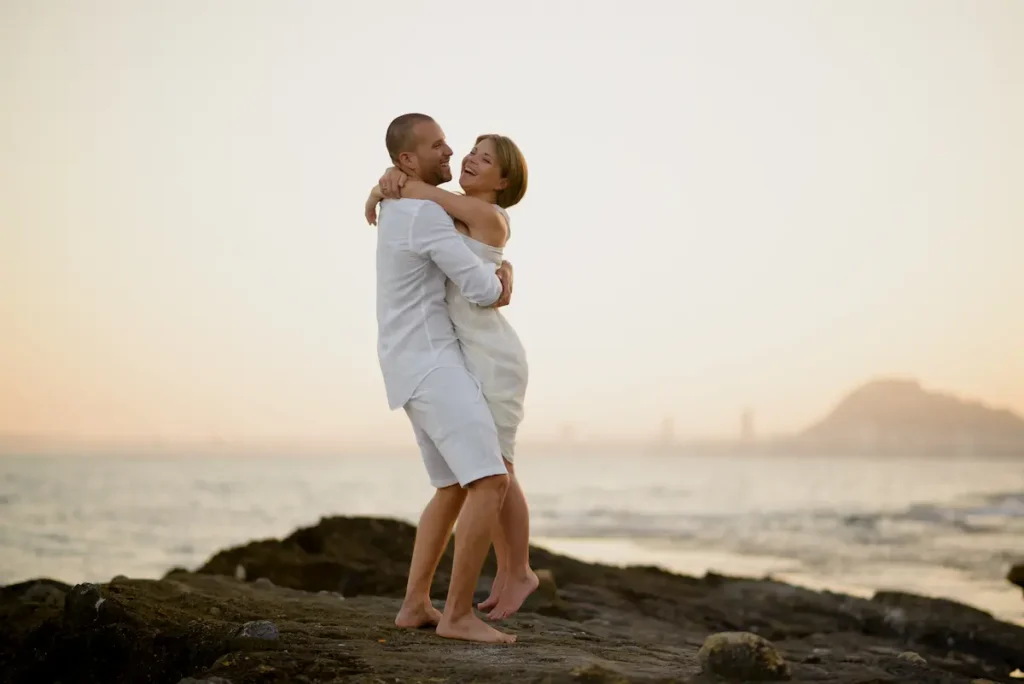 Couple embracing and smiling on a rocky beach at sunset, dressed in white.