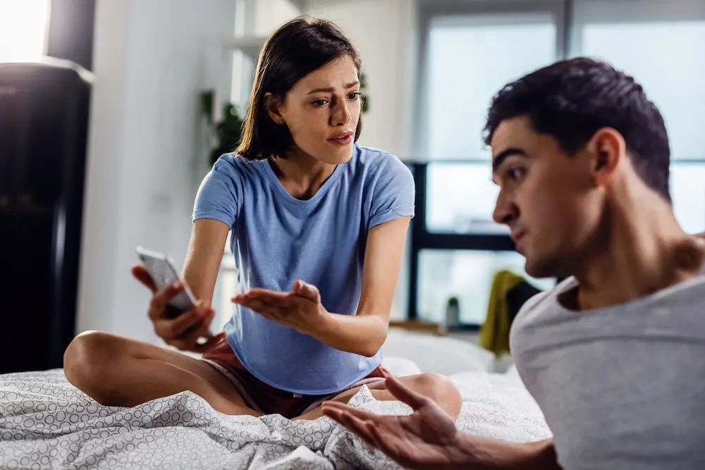 Couple arguing on a bed - Finding Peace Counseling Service PLLC - Finding Peace Counseling Service PLLC Couple arguing on a bed as the woman holds a phone, illustrating the emotional impact of infidelity.