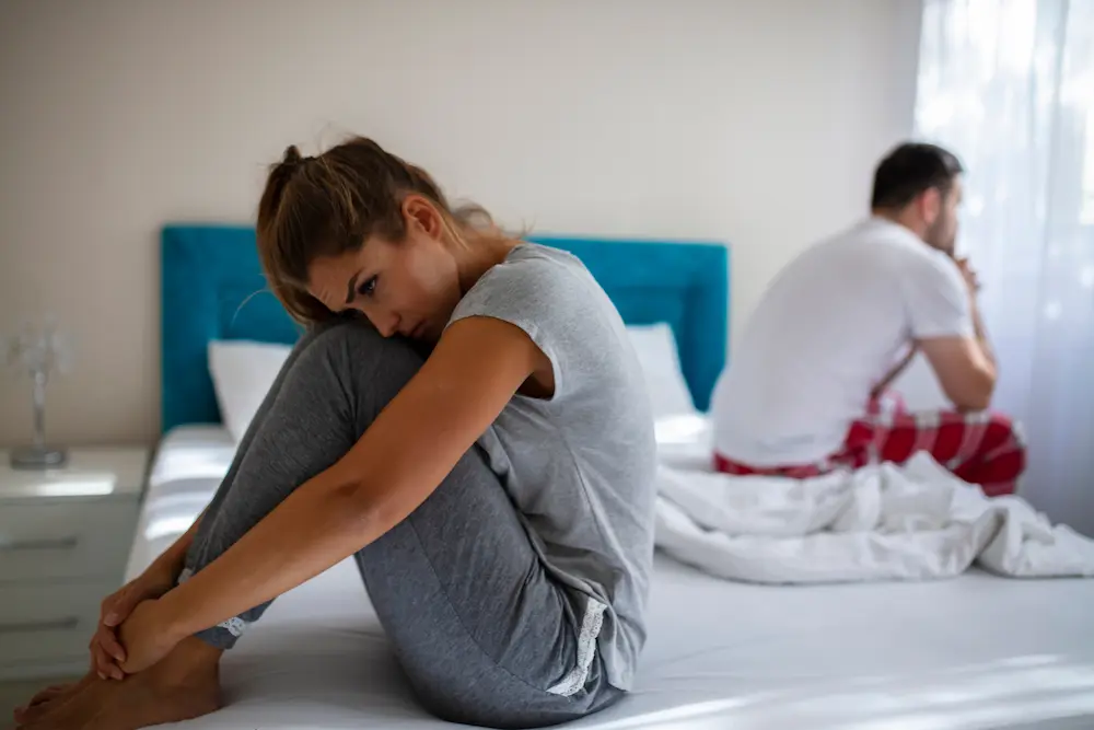 Woman sitting alone on a bed while her partner faces away in the background, depicting the emotional strain of infidelity.