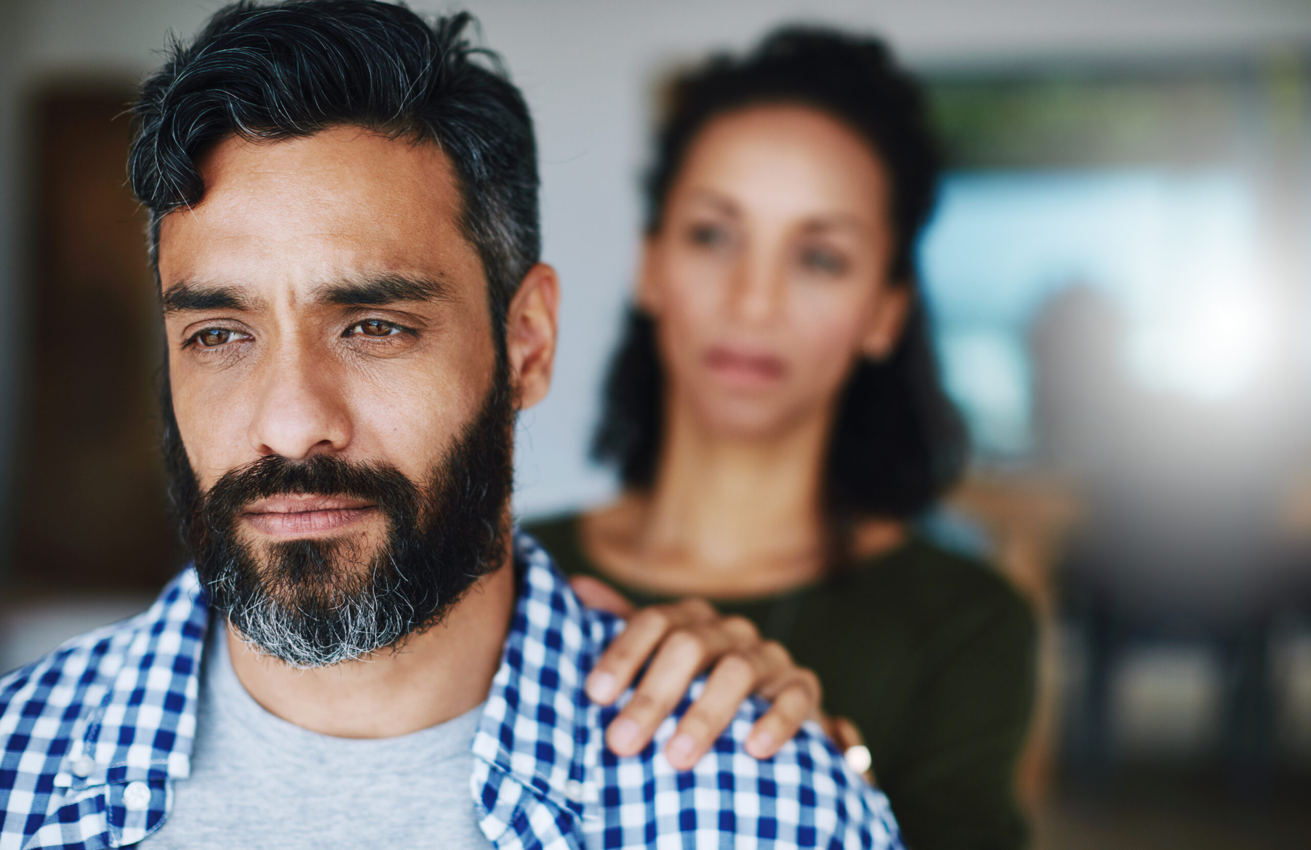 Man with dark hair and beard in a blue plaid shirt; a woman with a hand on his shoulder is blurred in the background.