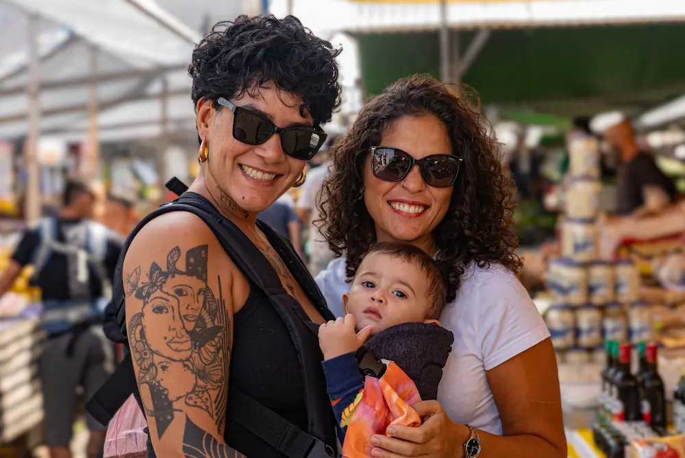 Two women at an outdoor market smiling with a baby in a carrier between them, sunny day and market stalls in the background.